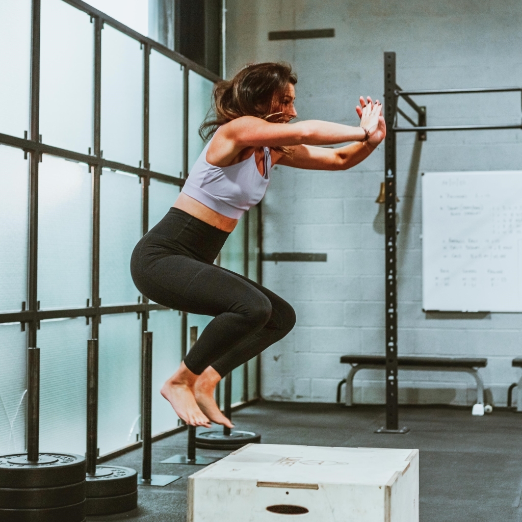 A woman mid-air performing a box jump in a gym. She is wearing a grey crop top and black leggings. Behind her are a whiteboard and gym equipment, including a pull-up bar. The gym has large frosted windows on one side.