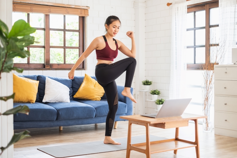 A woman in athletic wear exercises in a bright living room, lifting her knee as she follows a workout on a laptop. The room features a blue sofa with white and yellow cushions and a wooden coffee table.