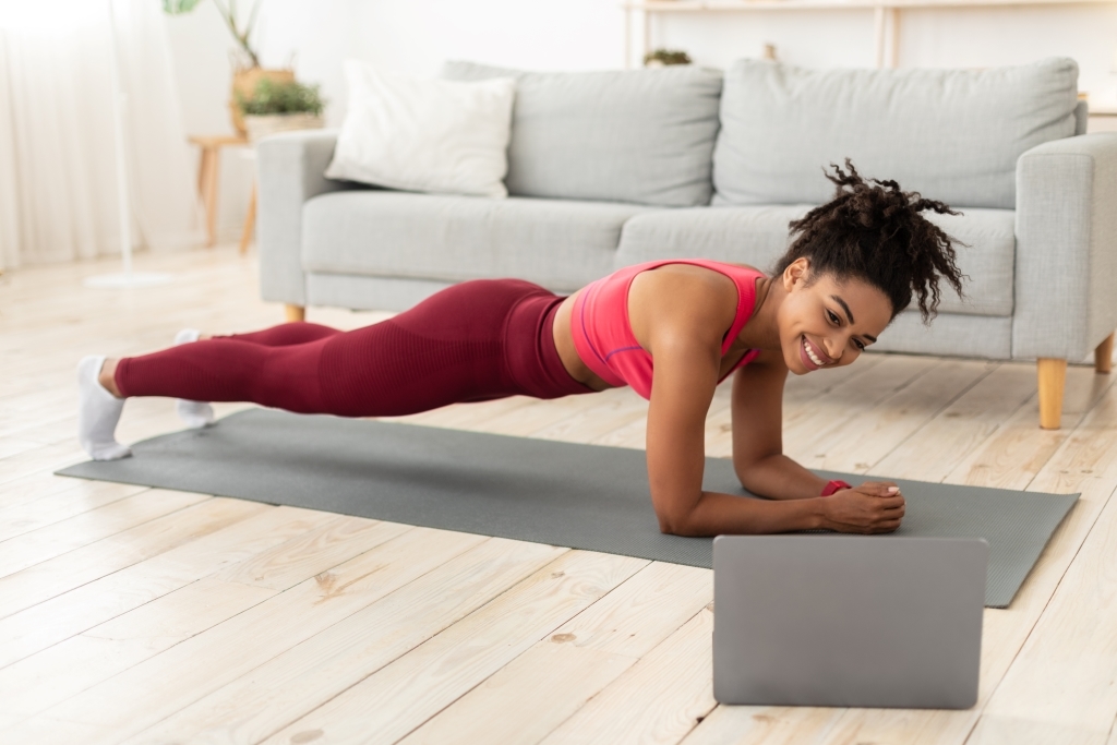 A woman is planking on a yoga mat in her living room, wearing a pink sports bra and maroon leggings. She is smiling and looking at a laptop on the floor. A gray couch and plants are in the background.